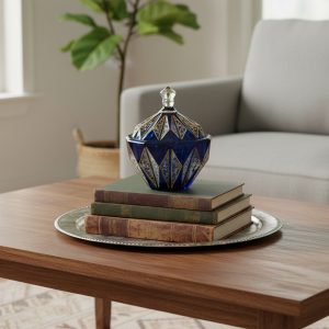 قندان مودنا ترمه (Modena Termeh sugar bowl) – A deep cobalt blue glass lidded bowl with intricate gold geometric and floral detailing, sitting atop a stack of vintage books on a wooden coffee table in a modern living room setting.