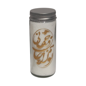 "White candle in a tall glass jar with a lit flame and prominent golden calligraphy design, on a dark brown table.")